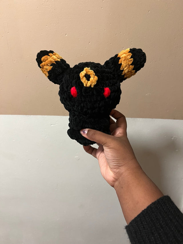 Front view of a handmade black chenille Umbreon crochet plush with gold ear bands and forehead ring, red embroidered eyes, and tiny tucked paws, held in one hand. Pattern by ElegantPurely.